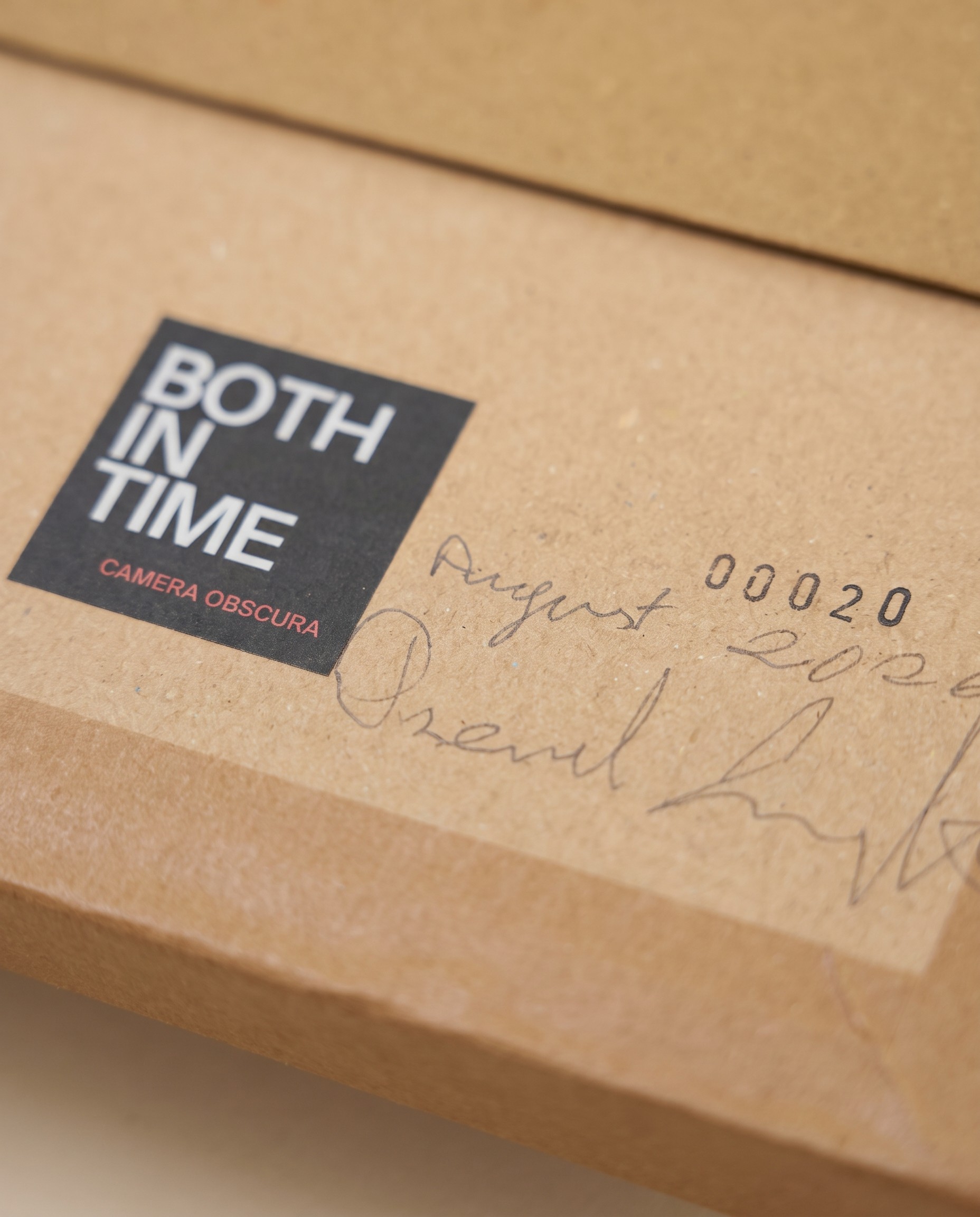 Nahaufnahme einer versiegelten Both In Time Kamera aus Karton: schwarzes Logo-Etikett, gestempelte Nummer 00020, daneben ein handschriftliches Datum und eine Unterschrift in Bleistift.